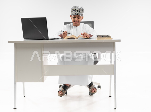 التعليم الالكتروني المنزلي، مدارس وأكاديميات متطوره، اتباع طرق تعليمية جديدة، التقنيه والتكنولوجيا الحديثه، بورتريه لصبي عربي خليجي عماني  يرتدي الدشداشة والكمة يقوم بمراجعة الدروس باستخدام الحاسوب المحمول، صورة لكامل الجسم، خلفية بيضاء
