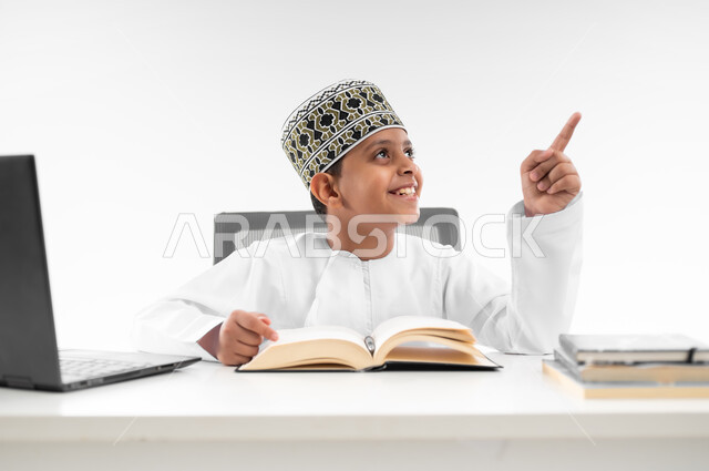 مراجعة الدروس باستخدام الحاسوب المحمول، مدارس وأكاديميات متطوره، اتباع طرق تعليمية جديدة، التقنيه والتكنولوجيا الحديثه، التعليم الالكتروني المنزلي، بورتريه مقرب لصبي عربي خليجي عماني  يرتدي الدشداشة والكمة يشير باصبعه لمكان ما، خلفية بيضاء