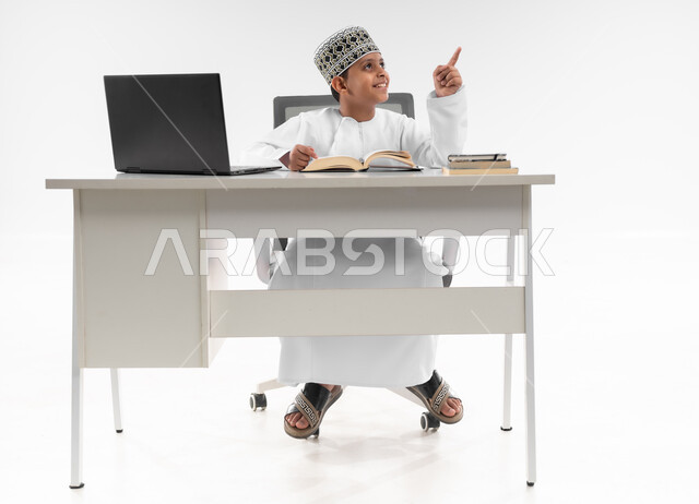 مراجعة الدروس باستخدام الحاسوب المحمول، مدارس وأكاديميات متطوره، اتباع طرق تعليمية جديدة، التقنيه والتكنولوجيا الحديثه، التعليم الالكتروني المنزلي، بورتريه لصبي عربي خليجي عماني  يرتدي الدشداشة والكمة يشير باصبعه لمكان ما، صورة لكامل الجسم، خلفية بيضاء