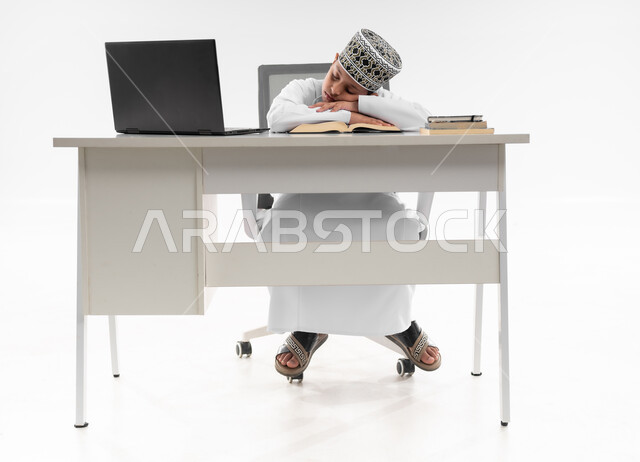 ايماءات النعاس والتعب والإرهاق، النوم على طاولة الدراسة، سلبيات التعلم عن بعد، هدر وضياع الوقت، استخدام التقنيات الحديثة، بورتريه لصبي عربي خليجي عماني  يرتدي الدشداشة والكمة يقوم بإهمال دروسها، صورة لكامل الجسم، خلفية بيضاء