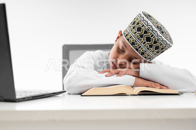 النوم على طاولة الدراسة، ايماءات النعاس والتعب والإرهاق، سلبيات التعلم عن بعد، هدر وضياع الوقت، استخدام التقنيات الحديثة، بورتريه مقرب  لصبي عربي خليجي عماني  يرتدي الدشداشة والكمة يقوم بإهمال دروسها، خلفية بيضاء