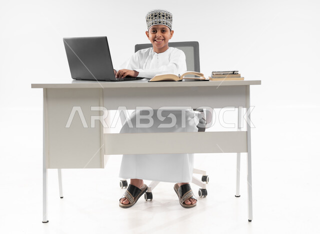 اتباع طرق تعليمية جديدة، مدارس وأكاديميات متطوره، استعمال التقنيه والتكنولوجيا الحديثه، التعليم الالكتروني المنزلي، بورتريه لصبي عربي خليجي عماني  يرتدي الدشداشة والكمة يقوم بمراجعة دروسه باستخدام الحاسوب المحمول، صورة لكامل الجسم، خلفية بيضاء