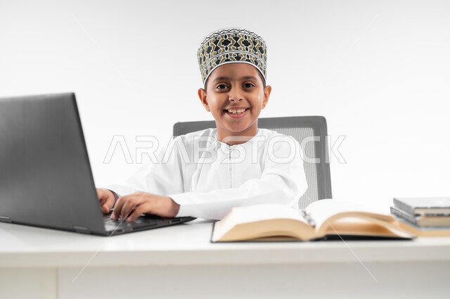 Looking at the camera with gestures of joy, following new educational methods, advanced schools and academies, using modern technology and technology, home e-learning, close-up portrait of an Arab Gulf Omani boy wearing a dishdasha and a turban reviewing his lessons using a laptop, white background
