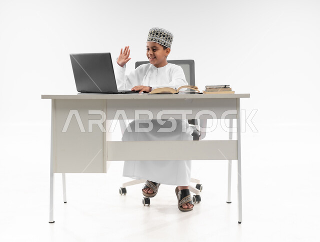 التعليم الالكتروني المنزلي، المشاركة الفعالة للطلاب ومتابعة الواجبات من خلال منصة التعلم عن بعد، متابعة الحصص وحل الواجبات عبر الانترنت، بورتريه لصبي عربي خليجي عماني  يرتدي الدشداشة والكمة يقوم بمراجعة دروسه باستخدام الحاسوب المحمول، صورة لكامل الجسم، خلفية بيضاء