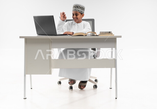 Active student participation and follow-up of assignments through the distance learning platform, home e-learning, follow-up of classes and solving assignments online, portrait of an Arab Gulf Omani boy wearing a dishdasha and a kummah reviewing his lessons using a laptop, full body image, white background