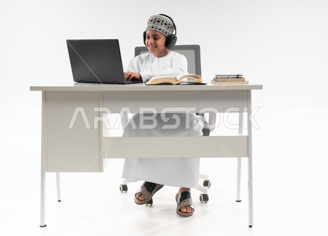مفهوم التعليم الإلكتروني، استخدام الاجهزة التقنية الحديثة، متابعة الحصص و حل الواجبات عبر الانترنت، بورتريه لصبي عربي خليجي عماني  يرتدي الدشداشة والكمة يضع سماعة الرأس يقوم بمراجعة دروسه، المذاكرة والدراسة عن بعد، صورة لكامل الجسم، خلفية بيضاء