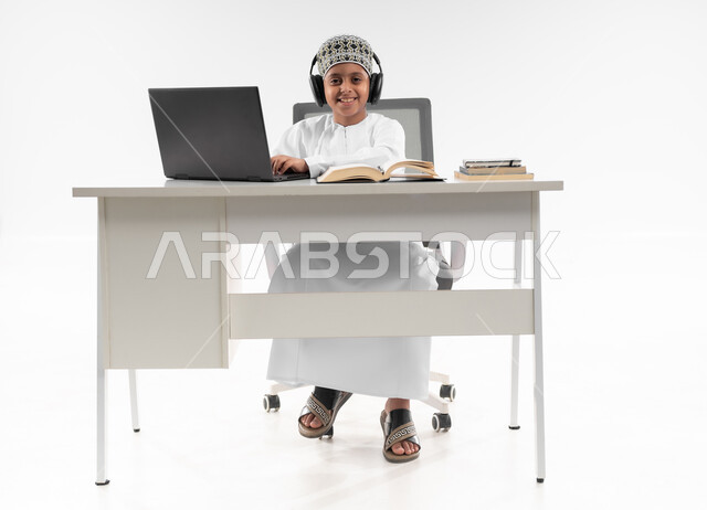 المذاكرة والدراسة عن بعد، استخدام الاجهزة التقنية الحديثة، مفهوم التعليم الإلكتروني، متابعة الحصص و حل الواجبات عبر الانترنت، بورتريه لصبي عربي خليجي عماني  يرتدي الدشداشة والكمة يضع سماعة الرأس يقوم بمراجعة دروسه، صورة لكامل الجسم، خلفية بيضاء