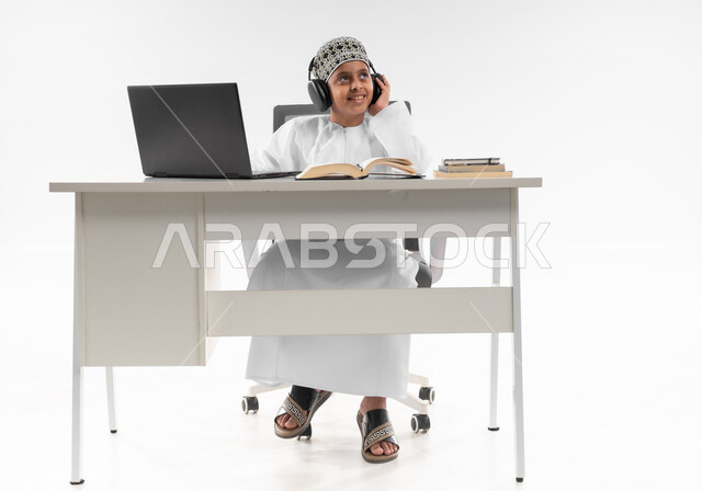 Using modern technical devices, the concept of e-learning, following up on classes and solving homework online, a portrait of an Arab Gulf Omani boy wearing a dishdasha and a kuma, wearing headphones, reviewing his lessons, studying and studying remotely, a full body image, white background