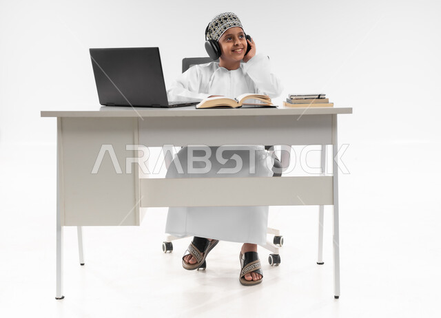 مفهوم التعليم الإلكتروني، استخدام الاجهزة التقنية الحديثة، متابعة الحصص و حل الواجبات عبر الانترنت، بورتريه لصبي عربي خليجي عماني  يرتدي الدشداشة والكمة يضع سماعة الرأس يقوم بمراجعة دروسه، المذاكرة والدراسة عن بعد، صورة لكامل الجسم، خلفية بيضاء