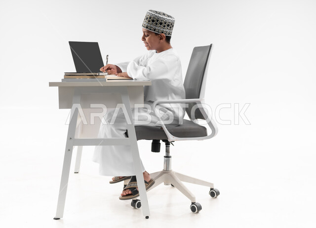 متابعة الحصص و حل الواجبات عبر الانترنت، استخدام الاجهزة التقنية الحديثة، بورتريه من الجانب لصبي عربي خليجي عماني  يرتدي الدشداشة والكمة يقوم بمراجعة دروسه، المذاكرة والدراسة عن بعد، مفهوم التعليم الإلكتروني، صورة لكامل الجسم، خلفية بيضاء