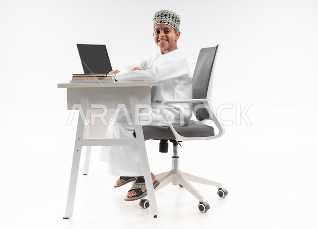 استخدام الاجهزة التقنية الحديثة، متابعة الحصص و حل الواجبات عبر الانترنت، بورتريه من الجانب لصبي عربي خليجي عماني  يرتدي الدشداشة والكمة يقوم بمراجعة دروسه، المذاكرة والدراسة عن بعد، مفهوم التعليم الإلكتروني، صورة لكامل الجسم، خلفية بيضاء