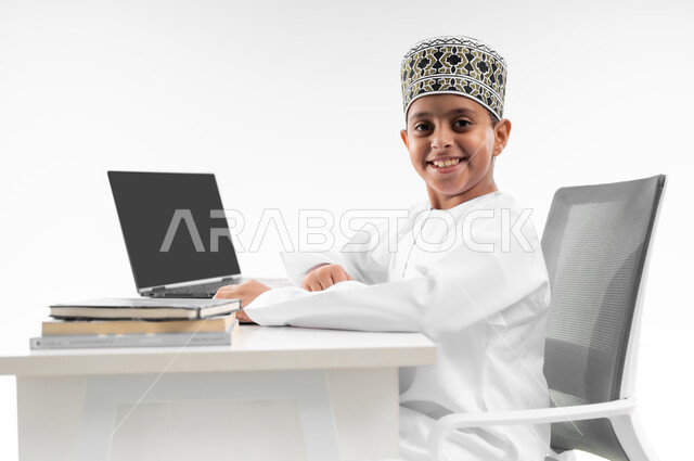 النظر الى الكاميرا بايماءات السعادة، استخدام الاجهزة التقنية الحديثة، متابعة الحصص و حل الواجبات عبر الانترنت، بورتريه مقرب من الجانب لصبي عربي خليجي عماني  يرتدي الدشداشة والكمة يقوم بمراجعة دروسه، المذاكرة والدراسة عن بعد، مفهوم التعليم الإلكتروني، خلفية بيضاء