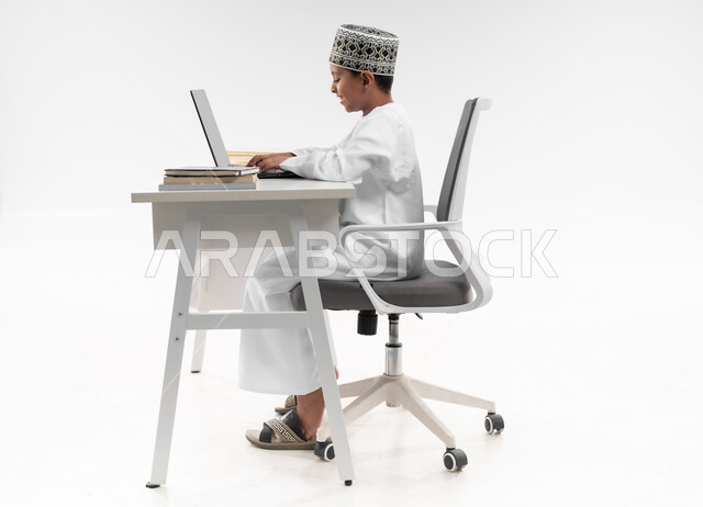 Following classes and solving homework online, using modern technical devices, side portrait of an Arab Gulf Omani boy wearing a dishdasha and a kimono reviewing his lessons, studying and distance learning, the concept of e-learning, a full body image, white background