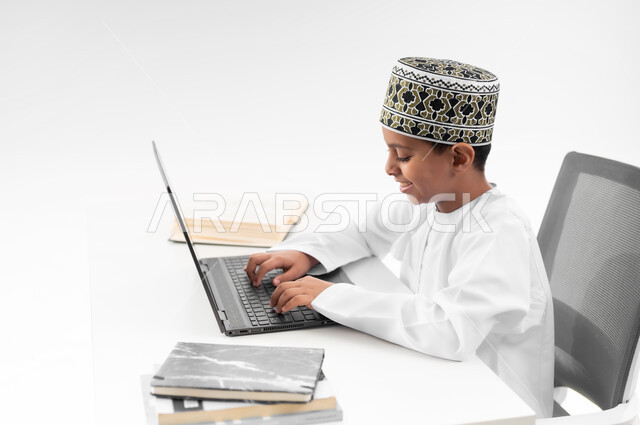 مفهوم التعليم الإلكتروني، استخدام الاجهزة التقنية الحديثة، متابعة الحصص و حل الواجبات عبر الانترنت، بورتريه مقرب من الجانب لصبي عربي خليجي عماني  يرتدي الدشداشة والكمة يقوم بمراجعة دروسه، المذاكرة والدراسة عن بعد، خلفية بيضاء
