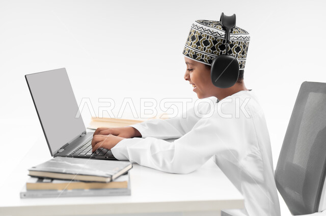 Studying and distance learning, the concept of e-learning, using modern technical devices, following classes and solving homework via the Internet, a close-up portrait from the side of an Arab Gulf Omani boy wearing a dishdasha and a cloak, wearing headphones, reviewing his lessons, white background