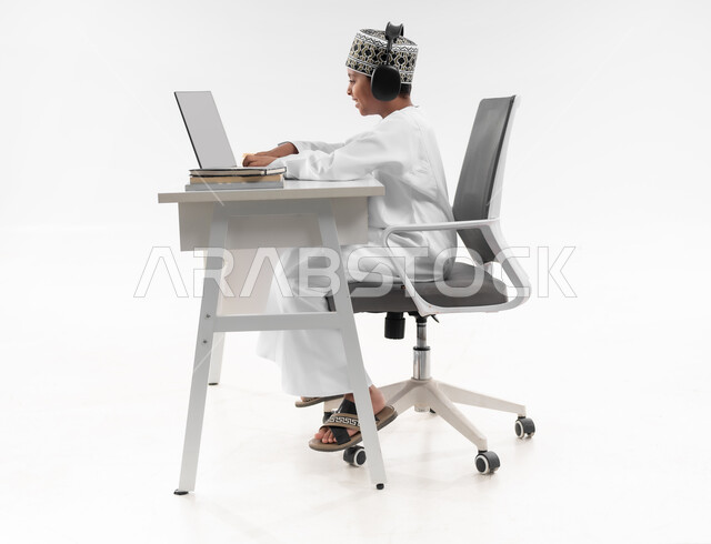 Preparing for exams and final tests, following classes and solving homework online, studying and studying remotely, the concept of e-learning, side portrait of an Arab Gulf Omani boy wearing a dishdasha and a turban, wearing headphones, reviewing his lessons, full body image, white background