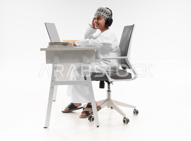 Solving homework online, studying and studying remotely, the concept of e-learning, using modern technical devices, a side portrait of an Arab Gulf Omani boy wearing a dishdasha and a kuma, wearing headphones, reviewing his lessons, a full body image, white background