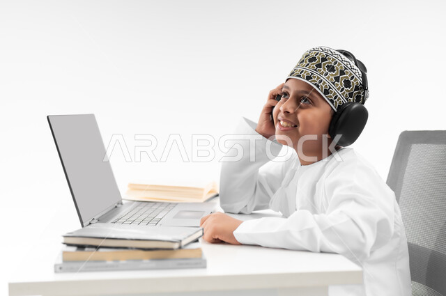 The concept of e-learning, using modern technical devices, following up on classes and solving homework online, a close-up portrait from the side of an Arab Gulf Omani boy wearing a dishdasha and a turban, wearing headphones, reviewing his lessons, studying and studying remotely, white background