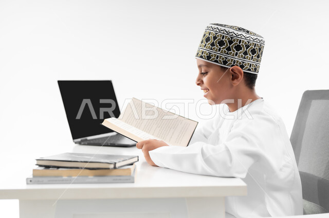 استخدام الاجهزة التقنية الحديثة، متابعة الحصص و حل الواجبات عبر الانترنت، بورتريه مقرب من الجانب لصبي عربي خليجي عماني  يرتدي الدشداشة والكمة يقوم بمراجعة دروسه، المذاكرة والدراسة عن بعد، مفهوم التعليم الإلكتروني، خلفية بيضاء