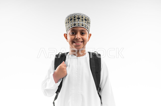البهجة بالعودة إلى المدارس، الاستعداد للعام الدراسي الجديد، التعليم في سلطنة عمان، بورتريه مقرب لتلميذ عربي خليجي عماني يرتدي الدشداشة والكمة يحمل الحقيبة المدرسية ينظر الى الكاميرا بايماءات السرور والسعادة، خلفيه بيضاء
