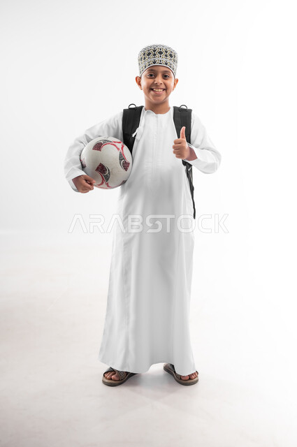 لعب الحصص الرياضية الممتعة، جمع التعلم مع الترفيه، بورتريه لتلميذ عربي خليجي عماني يرتدي الدشداشة والكمة يحمل الحقيبة المدرسية يمسك بيده كرة القدم يرفع اصبع الابهام بايماءات القبول والموافقة، صورة لكامل الجسم، خلفيه بيضاء