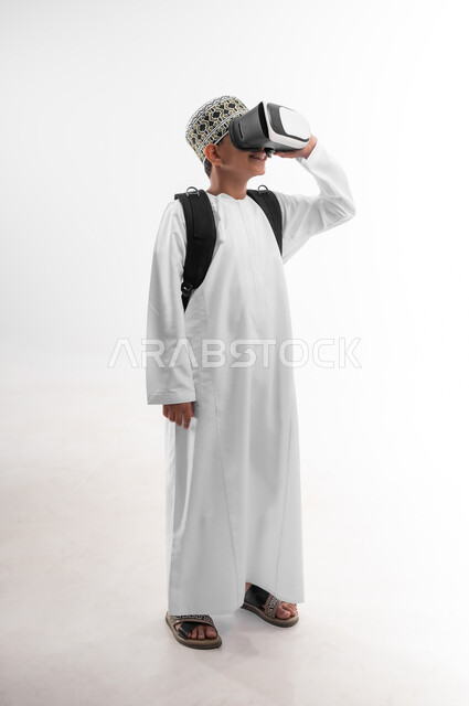 جمع التعلم مع الترفيه، مدارس وأكاديميات متطوره، اتباع طرق تعليمية جديدة، بورتريه من الجانب لتلميذ عربي خليجي عماني يرتدي الدشداشة والكمة يحمل الحقيبة المدرسية والكتاب يضع نظارات الواقع الافتراضي، استخدام التقنيه والتكنولوجيا الحديثه، صورة لكامل الجسم، خلفيه بيضاء