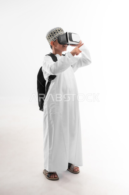 Using technology and modern technology, combining learning with entertainment, advanced schools and academies, side portrait of an Arab Gulf Omani student wearing a dishdasha and a turban, carrying a school bag and a book, wearing virtual reality glasses, pointing to a place, full body image, white background