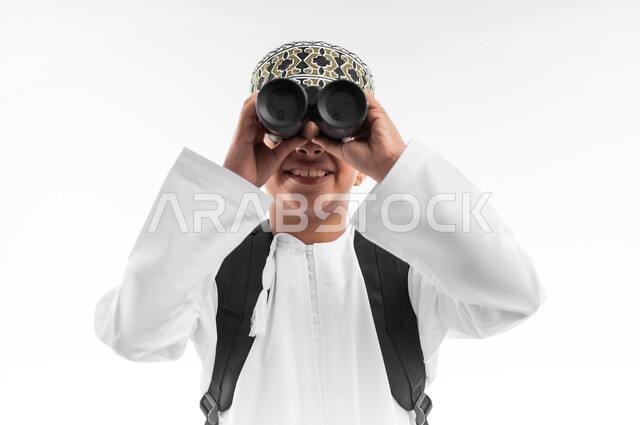 رؤية الأشياء البعيدة باستخدام التلسكوب، حب الاستكشاف والمغامرة، بورتريه مقرب لطالب عربي خليجي عماني يرتدي الدشداشة والكمة يحمل حقيبة الظهر المدرسية ينظر من خلال المنظار، التخطيط المستقبلي للأهداف والطموحات، الخلفية البيضاء
