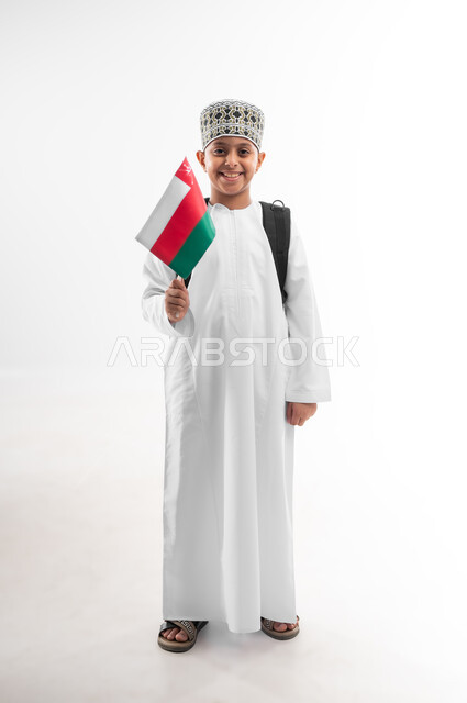 الأعلام الوطنية للدول العربية، الأعياد والمناسبات الوطنية، الفخر والاعتزاز بالرموز والراية العمانية، العودة الى المدارس، بورتريه لطالب عربي خليجي عماني  يرتدي الدشداشة والكمة يحمل حقيبة الظهر المدرسية يمسك بيده علم سلطنة عمان، صورة لكامل الجسم، خلفية بيضاء