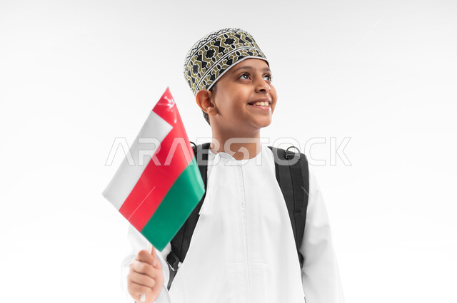الرايات والأعلام الوطنية للدول العربية، الأعياد والمناسبات الوطنية، الفخر والاعتزاز بالرموز والراية العمانية، العودة الى المدارس، بورتريه مقرب لطالب عربي خليجي عماني  يرتدي الدشداشة والكمة يحمل حقيبة الظهر المدرسية يمسك بيده علم سلطنة عمان، خلفية بيضاء