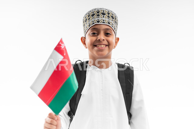 المشاركة بالأعياد والمناسبات الوطنية، الفخر والاعتزاز بالرموز والراية العمانية، العودة الى المدارس، بورتريه مقرب لطالب عربي خليجي عماني  يرتدي الدشداشة والكمة يحمل حقيبة الظهر المدرسية يمسك بيده علم سلطنة عمان، خلفية بيضاء