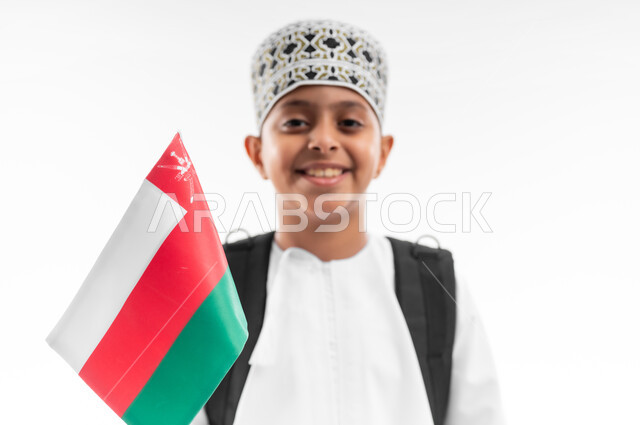 Pride and appreciation for Omani symbols and flag, participation in national holidays and occasions, back to school, close-up portrait of an Arab Gulf Omani student wearing a dishdasha and a kummah carrying a school backpack holding the flag of the Sultanate of Oman, white background