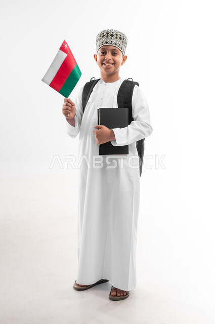 الأعياد والمناسبات الوطنية، الفخر والاعتزاز بالرموز والراية العمانية، العودة الى المدارس، بورتريه لطالب عربي خليجي عماني  يرتدي الدشداشة والكمة يحمل حقيبة الظهر المدرسية يمسك بيده الكتاب وعلم سلطنة عمان، صورة لكامل الجسم، خلفية بيضاء