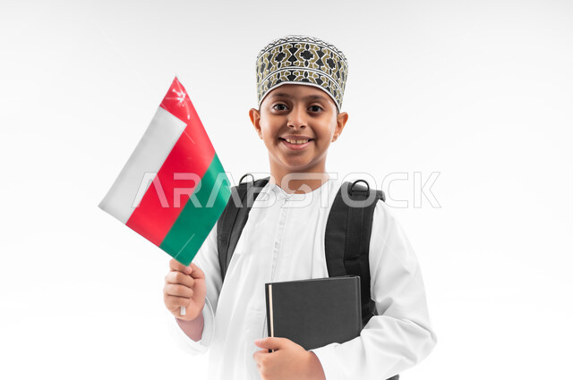 المشاركة بالأعياد والمناسبات الوطنية، الفخر والاعتزاز بالرموز والراية العمانية، العودة الى المدارس، بورتريه مقرب لطالب عربي خليجي عماني  يرتدي الدشداشة والكمة يحمل حقيبة الظهر المدرسية يمسك بيده الكتاب وعلم سلطنة عمان، خلفية بيضاء