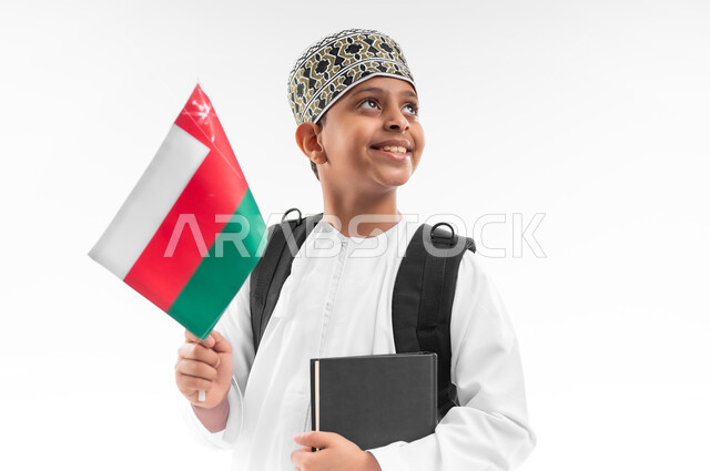 الفخر والاعتزاز بالرموز والراية العمانية، المشاركة بالأعياد والمناسبات الوطنية، العودة الى المدارس، بورتريه مقرب لطالب عربي خليجي عماني  يرتدي الدشداشة والكمة يحمل حقيبة الظهر المدرسية يمسك بيده الكتاب وعلم سلطنة عمان، خلفية بيضاء