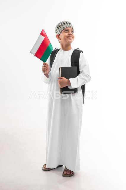 الرايات والأعلام الوطنية للدول العربية، الفخر والاعتزاز بالرموز والراية العمانية، العودة الى المدارس، بورتريه لطالب عربي خليجي عماني  يرتدي الدشداشة والكمة يحمل حقيبة الظهر المدرسية يمسك بيده الكتاب وعلم سلطنة عمان، صورة لكامل الجسم، خلفية بيضاء
