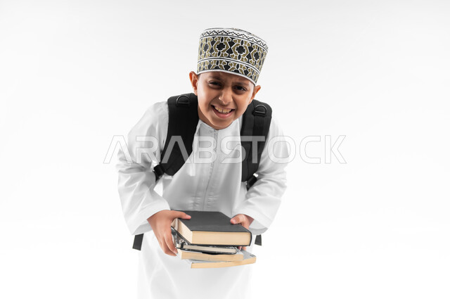 العودة الى المدارس، المناهج العلمية التعليمية، مفهوم الجد والاجتهاد للوصول إلى النجاح، مراجعة الدروس وحل الواجبات، بورتريهمقرب لطالب عربي خليجي عماني  يرتدي الدشداشة والكمة يحمل حقيبة الظهر المدرسية يمسك بيده مجموعة من الكتب بايماءات التعب، خلفية بيضاء