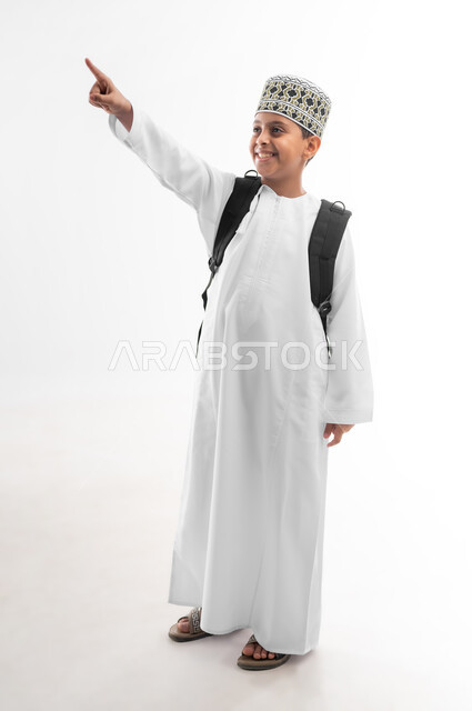Pointing to a specific location, back to school concept, educational scientific curricula, portrait of an Arab Gulf Omani student wearing a dishdasha and a kummah carrying a school backpack pointing to a place, full body image, white background