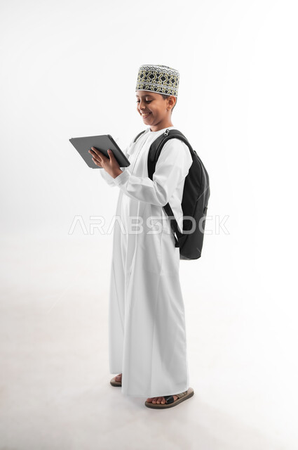 Following classes and solving homework online, using modern technical devices, studying and distance learning, side portrait of an Arab Gulf Omani student wearing a dishdasha and a turban reviewing lessons using a tablet, e-learning concept, full body image, white background