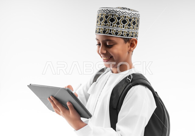 استخدام الاجهزة التقنية الحديثة، المذاكرة والدراسة عن بعد، متابعة الحصص و حل الواجبات عبر الانترنت، بورتريه مقرب من الجانب لطالب عربي خليجي عماني يرتدي الدشداشة والكمة يقوم بمراجعة الدروس باستعمال التابلت، مفهوم التعليم الإلكتروني، خلفية بيضاء