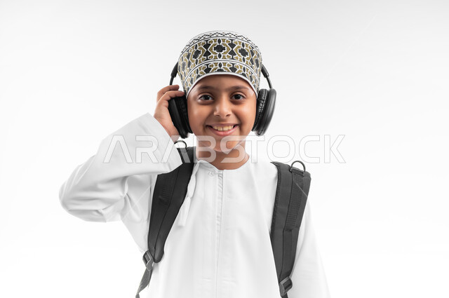 Looking at the camera with gestures of pleasure, educational scientific curricula, the concept of back to school, seriousness and diligence to achieve success, close-up portrait of an Arab Gulf Omani student wearing a dishdasha and a cloak, wearing headphones, carrying a school backpack, white background