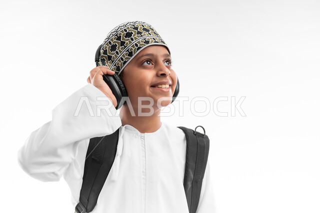 المناهج العلمية التعليمية، النظر الى مكان ما بايماءات السرور، مفهوم العودة إلى المدارس، الجد والاجتهاد للوصول إلى النجاح، بورتريه مقرب لطالب عربي خليجي عماني يرتدي الدشداشة والكمة يضع سماعة الرأس يحمل حقيبة الظهر المدرسية، خلفية بيضاء