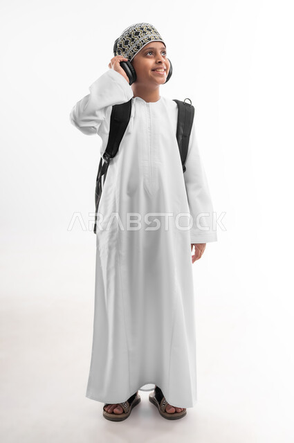 مفهوم العودة إلى المدارس، المناهج العلمية التعليمية، النظر الى مكان ما بايماءات السرور، الجد والاجتهاد للوصول إلى النجاح، بورتريه لطالب عربي خليجي عماني يرتدي الدشداشة والكمة يضع سماعة الرأس يحمل حقيبة الظهر المدرسية، صورة لكامل الجسم، خلفية بيضاء
