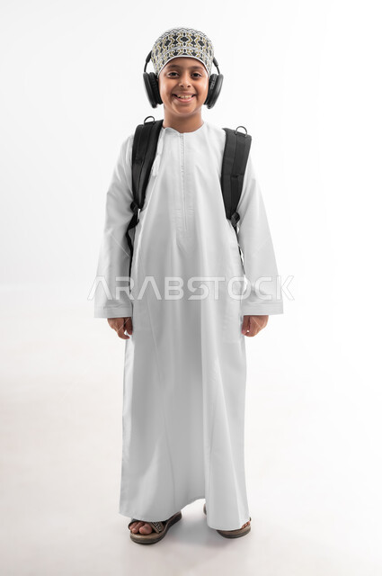 النظر الى الكاميرا بايماءات السرور، المناهج العلمية التعليمية، مفهوم العودة إلى المدارس، الجد والاجتهاد للوصول إلى النجاح، بورتريه لطالب عربي خليجي عماني يرتدي الدشداشة والكمة يضع سماعة الرأس يحمل حقيبة الظهر المدرسية، صورة لكامل الجسم، خلفية بيضاء