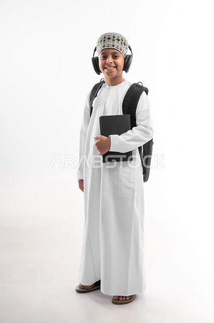 مفهوم الاجتهاد للوصول إلى النجاح، مراجعة الدروس وحل الواجبات، المناهج العلمية التعليمية، مفهوم العودة إلى المدارس، بورتريه لطالب عربي خليجي عماني يرتدي الدشداشة والكمة يضع سماعة الرأس يحمل حقيبة الظهر المدرسية يحمل بيده كتاب، صورة لكامل الجسم، خلفية بيضاء