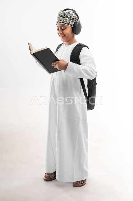مفهوم الاجتهاد للوصول إلى النجاح، مراجعة الدروس وحل الواجبات، المناهج العلمية التعليمية، مفهوم العودة إلى المدارس، بورتريه من الجانب لطالب عربي خليجي عماني يرتدي الدشداشة والكمة يضع سماعة الرأس يحمل حقيبة الظهر المدرسية يحمل بيده كتاب، صورة لكامل الجسم، خلفية بيضاء