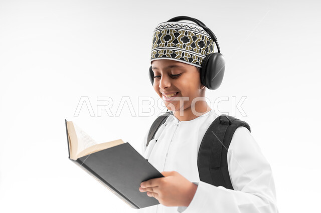 مراجعة الدروس وحل الواجبات، المناهج العلمية التعليمية، مفهوم العودة إلى المدارس، الجد والاجتهاد للوصول إلى النجاح، بورتريه مقرب من الجانب لطالب عربي خليجي عماني يرتدي الدشداشة والكمة يضع سماعة الرأس يحمل حقيبة الظهر المدرسية يحمل بيده كتاب، خلفية بيضاء