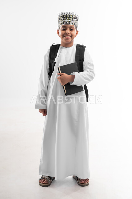 الوقوف باستقامة والنظر الى الكاميرا بايماءات السرور، المناهج العلمية التعليمية، مفهوم العودة إلى المدارس، الجد والاجتهاد للوصول إلى النجاح، بورتريه لطالب عربي خليجي عماني يرتدي الدشداشة والكمة يحمل حقيبة الظهر المدرسية يمسك بيده كتاب، صورة لكامل الجسم، خلفية بيضاء