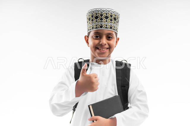 المناهج العلمية التعليمية، مفهوم العودة إلى المدارس، الجد والاجتهاد للوصول إلى النجاح، بورتريه مقرب لطالب عربي خليجي عماني يرتدي الدشداشة والكمة يحمل حقيبة الظهر المدرسية يمسك بيده كتاب يرفع اصبع الابهام بايماءات الاعجاب والرضى، خلفية بيضاء