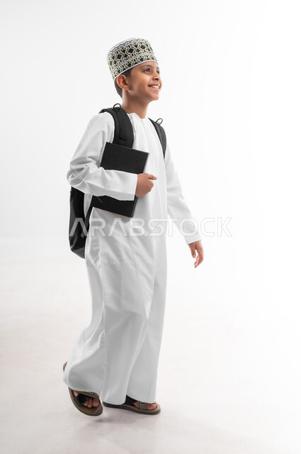 النظر الى مكان ما بإيماءات السعادة والفرح، الجد والاجتهاد للوصول إلى النجاح، مفهوم العودة إلى المدارس، بورتريه لطالب عربي خليجي عماني يرتدي الدشداشة والكمة يحمل حقيبة الظهر المدرسية يمسك بيده كتاب، المناهج العلمية التعليمية، صورة لكامل الجسم، خلفية بيضاء